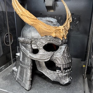 May include: A 3D printed helmet featuring a metallic silver skull-like face with sharp teeth and prominent fangs. The helmet is adorned with large, textured gold-colored horns extending upwards from the brow. The word CREALITY is visible.