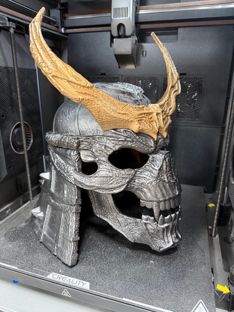 May include: A 3D printed helmet featuring a metallic silver skull-like face with sharp teeth and prominent fangs. The helmet is adorned with large, textured gold-colored horns extending upwards from the brow. The word CREALITY is visible.