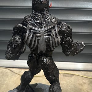 May include: A black Venom action figure with a white spider emblem on its back. The figure has a textured, glossy finish and a red tongue protruding from its mouth. The figure is posed on a gray base.