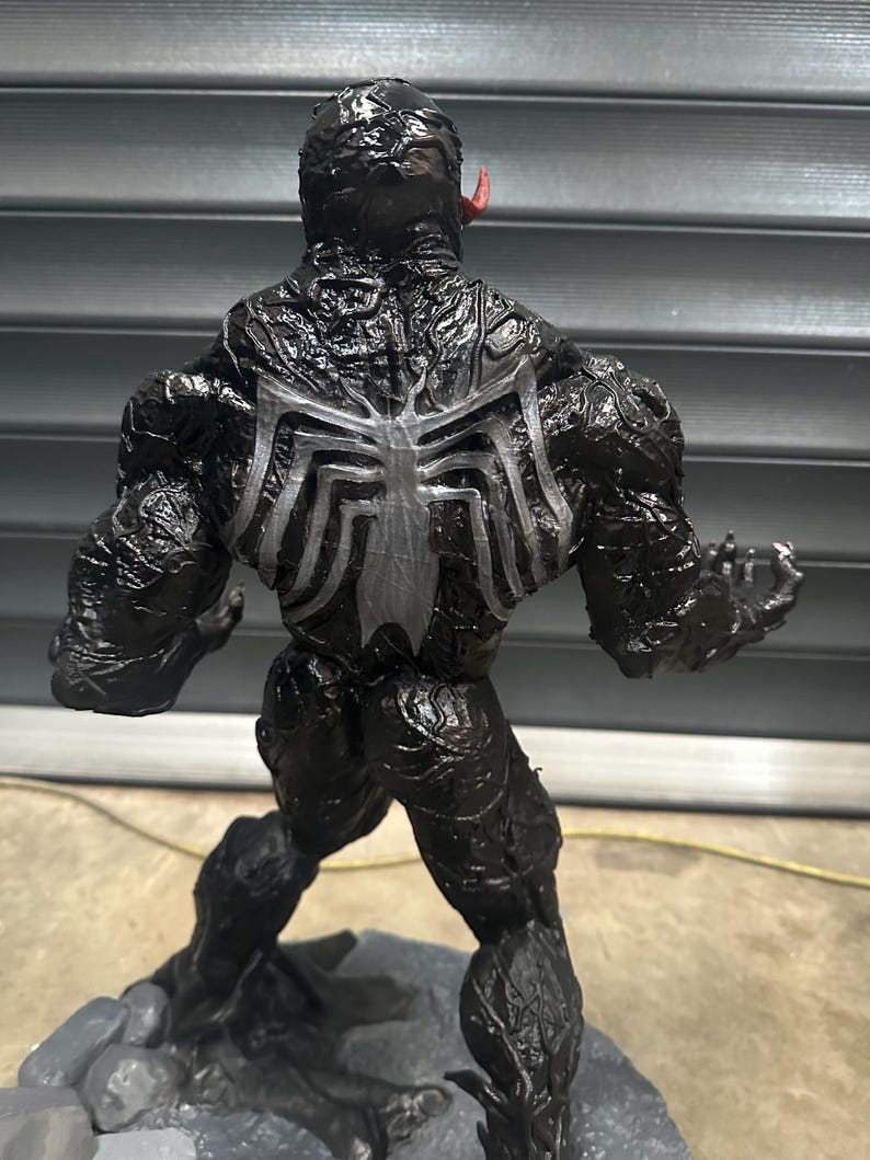 May include: A black Venom action figure with a white spider emblem on its back. The figure has a textured, glossy finish and a red tongue protruding from its mouth. The figure is posed on a gray base.