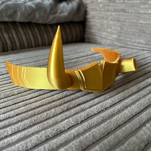 Puede incluir: Una corona dorada con una espiga central en forma de cuerno y detalles geom&eacute;tricos. La corona tiene una banda curva y probablemente est&aacute; dise&ntilde;ada para cosplay o disfraces. El material parece ser pl&aacute;stico impreso en 3D.