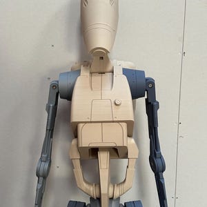 May include: A beige and gray plastic model of a robot with a humanoid shape. The robot has a large head with a rounded top and two small, circular eyes. The robot's arms are extended out to the sides, and its legs are bent at the knees.