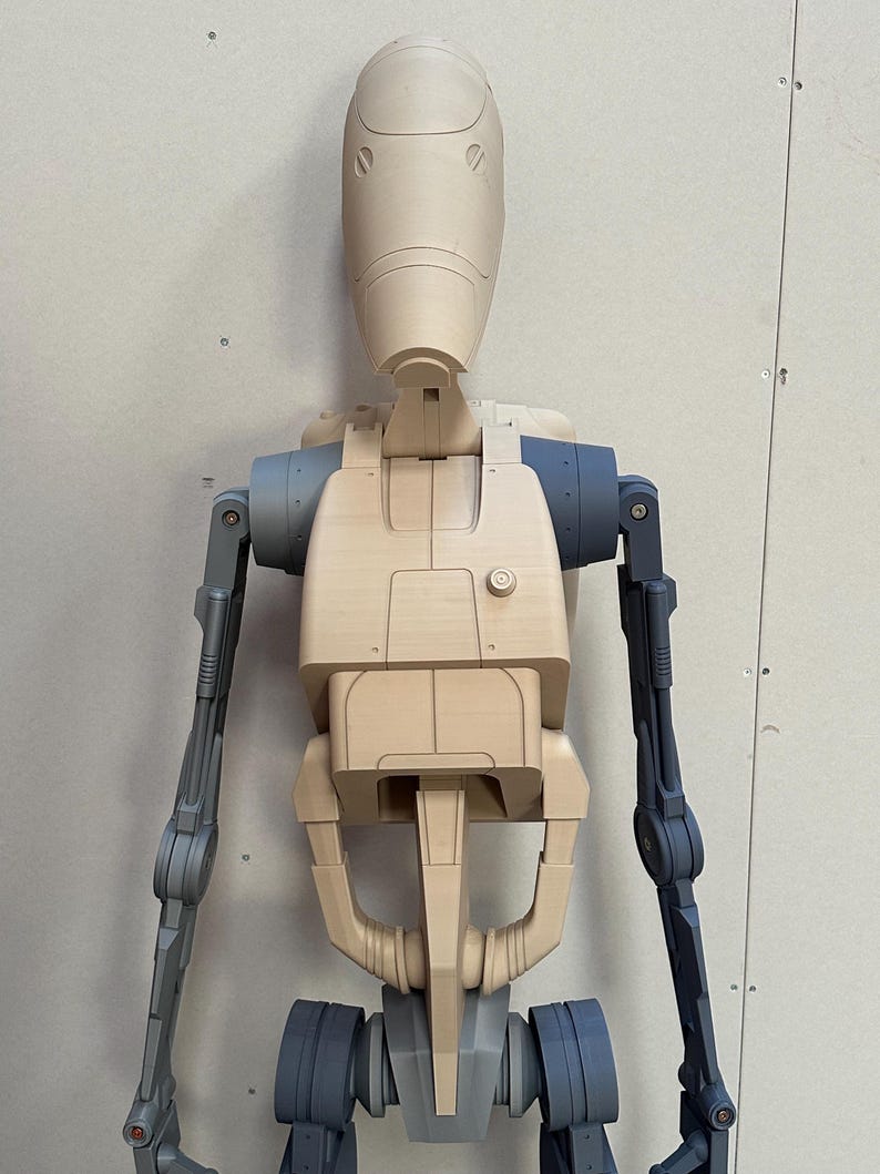 May include: A beige and gray plastic model of a robot with a humanoid shape. The robot has a large head with a rounded top and two small, circular eyes. The robot's arms are extended out to the sides, and its legs are bent at the knees.