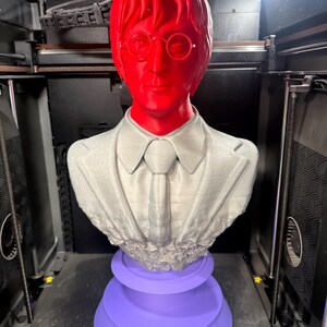 May include: A 3D-printed bust of a man with red hair and glasses, wearing a suit and tie. The bust is gray and rests on a purple, tiered base. The bust is in a 3D printer.