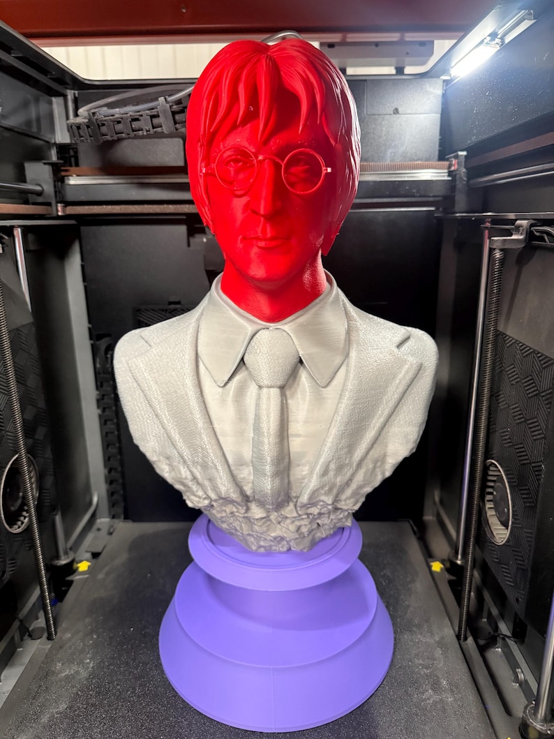 May include: A 3D-printed bust of a man with red hair and glasses, wearing a suit and tie. The bust is gray and rests on a purple, tiered base. The bust is in a 3D printer.