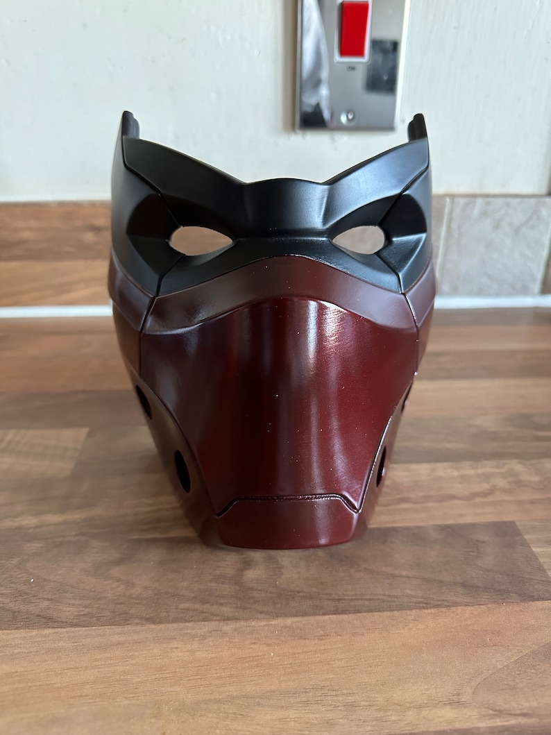 May include: A red and black plastic mask with a sleek, modern design. The mask has a raised, textured surface and two eye holes.