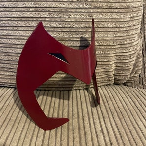 May include: A red plastic mask with a black design. The mask is shaped like a stylized face with a pointed chin and a wide forehead.