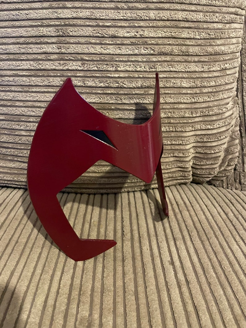 May include: A red plastic mask with a black design. The mask is shaped like a stylized face with a pointed chin and a wide forehead.