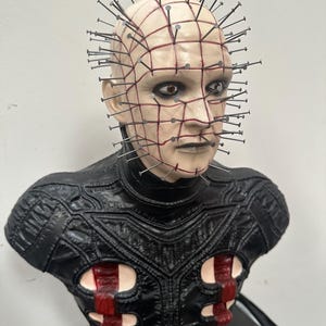 May include: A bust of a character with a pale face, grid-like markings, and numerous nails embedded in the head. The character has dark eyes and a stern expression. The bust is wearing a black leather-like garment with red accents.