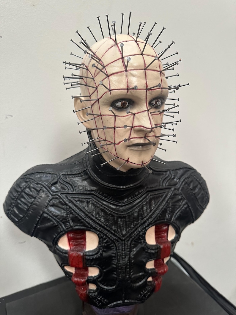 May include: A bust of a character with a pale face, grid-like markings, and numerous nails embedded in the head. The character has dark eyes and a stern expression. The bust is wearing a black leather-like garment with red accents.