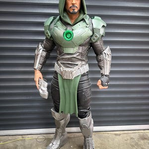 May include: A detailed action figure of a character in a green and silver armored suit with a hooded cloak. The figure has a stern expression and is holding a silver gauntlet. The figure is standing in front of a gray metal shutter.
