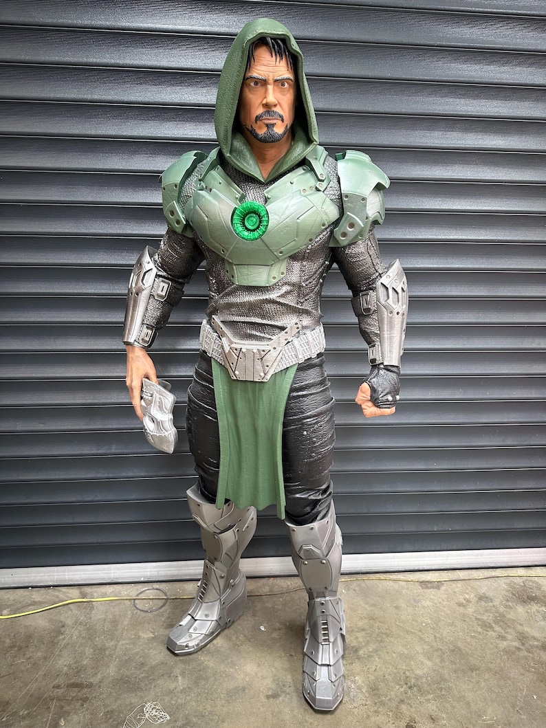 May include: A detailed action figure of a character in a green and silver armored suit with a hooded cloak. The figure has a stern expression and is holding a silver gauntlet. The figure is standing in front of a gray metal shutter.