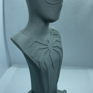 May include: A grey 3D printed bust of Spider-Man from the video game Spider-Man. The bust features a detailed sculpt of Spider-Man's head and torso, including his iconic webbed suit and spider symbol. The bust is mounted on a small base with the word "Spider-Man" printed on it.