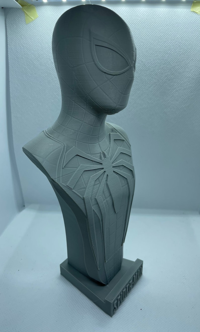 May include: A grey 3D printed bust of Spider-Man from the video game Spider-Man. The bust features a detailed sculpt of Spider-Man's head and torso, including his iconic webbed suit and spider symbol. The bust is mounted on a small base with the word "Spider-Man" printed on it.