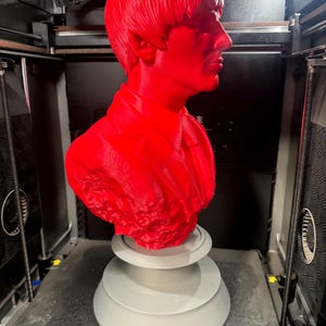 May include: A bright red bust of a man with a distinctive hairstyle, likely a celebrity, is displayed on a gray, tiered base. The bust is in the process of being 3D printed, with the printer visible in the background.