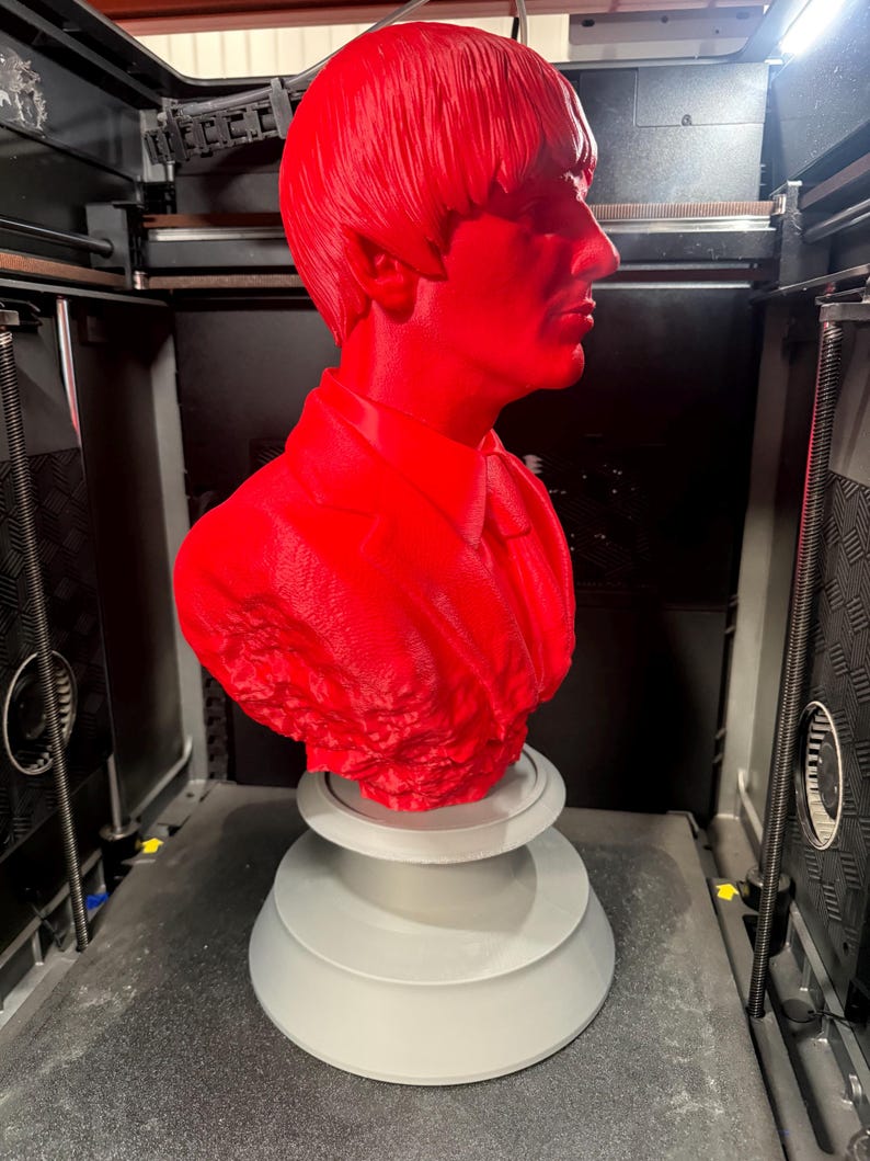 May include: A bright red bust of a man with a distinctive hairstyle, likely a celebrity, is displayed on a gray, tiered base. The bust is in the process of being 3D printed, with the printer visible in the background.