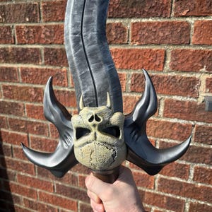 May include: A fantasy sword with a curved, gray blade and a detailed skull-shaped hilt. The hilt features curved, black accents and small horns. The handle is brown. The sword is held against a brick wall.