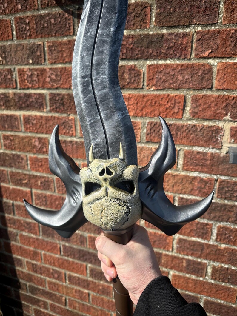 May include: A fantasy sword with a curved, gray blade and a detailed skull-shaped hilt. The hilt features curved, black accents and small horns. The handle is brown. The sword is held against a brick wall.