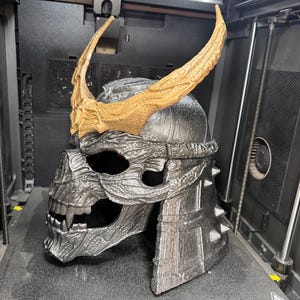 May include: A 3D printed helmet featuring a metallic silver skull design with a textured surface, sharp teeth, and prominent, curved, textured gold-colored horns attached to the forehead area. The helmet has a layered, segmented back section.
