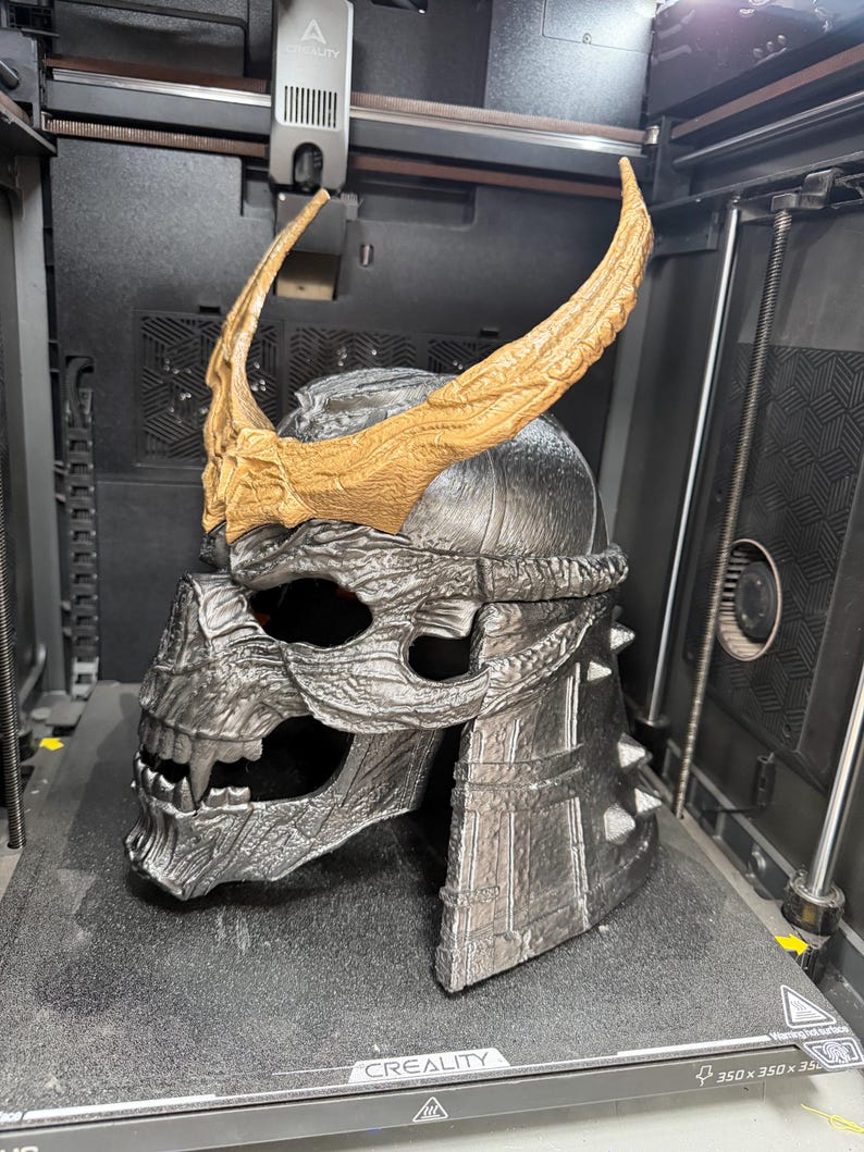 May include: A 3D printed helmet featuring a metallic silver skull design with a textured surface, sharp teeth, and prominent, curved, textured gold-colored horns attached to the forehead area. The helmet has a layered, segmented back section.