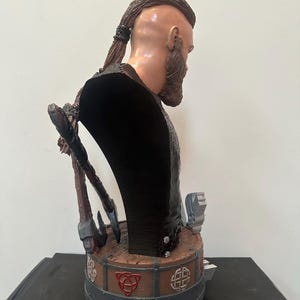 May include: A detailed bust of a Viking warrior, with a sculpted face, full beard, and braided hair. The figure wears a dark garment and is adorned with axes and Celtic knotwork. The bust is painted in realistic tones, showcasing intricate details.