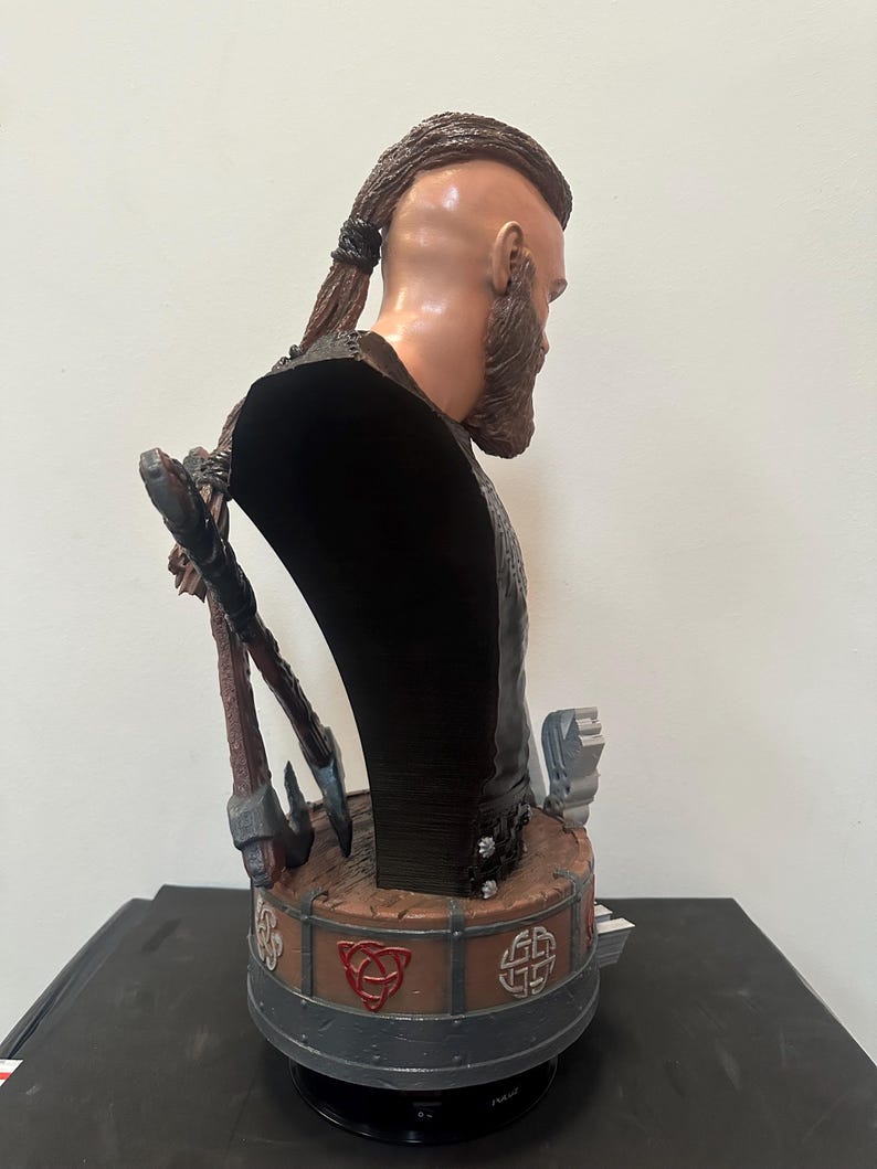 May include: A detailed bust of a Viking warrior, with a sculpted face, full beard, and braided hair. The figure wears a dark garment and is adorned with axes and Celtic knotwork. The bust is painted in realistic tones, showcasing intricate details.