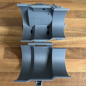 May include: A grey 3D printed object with a hinged lid and a small lever. The object is open and the inside is visible.