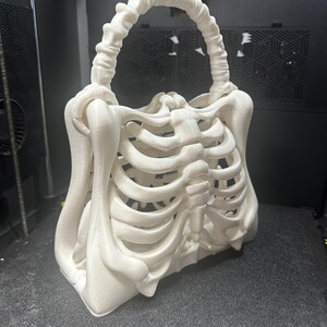 May include: A white, 3D-printed handbag in the shape of a human rib cage. The bag features a handle made of bone-like material and a rectangular base. The design is detailed, with visible ribs and sternum, creating a unique, gothic aesthetic.