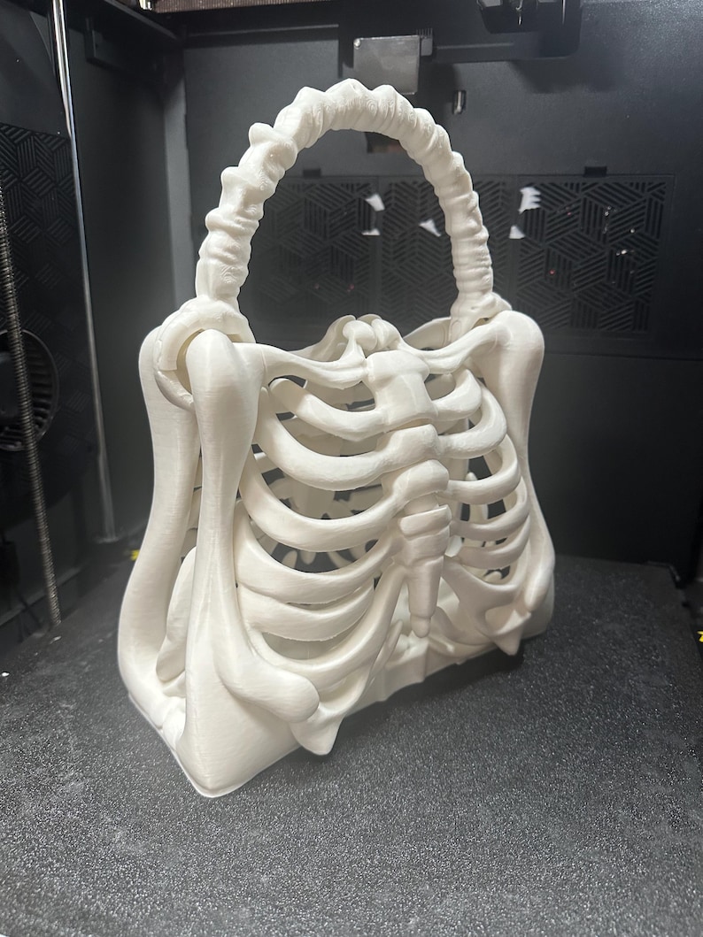 May include: A white, 3D-printed handbag in the shape of a human rib cage. The bag features a handle made of bone-like material and a rectangular base. The design is detailed, with visible ribs and sternum, creating a unique, gothic aesthetic.