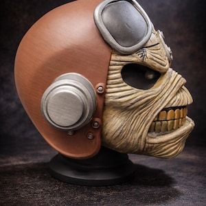 May include: A brown and tan sculpted bust of a skeletal face wearing a brown helmet with silver goggles and ear protection. The face has exposed teeth and is wrapped in bandages. The bust is mounted on a black base.