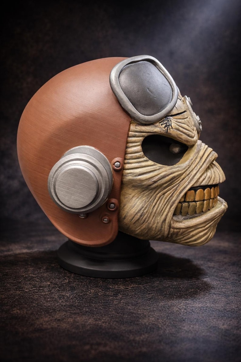 May include: A brown and tan sculpted bust of a skeletal face wearing a brown helmet with silver goggles and ear protection. The face has exposed teeth and is wrapped in bandages. The bust is mounted on a black base.