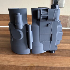 May include: A grey 3D printed model of a futuristic device with a gear on the side. The device is split into two pieces.