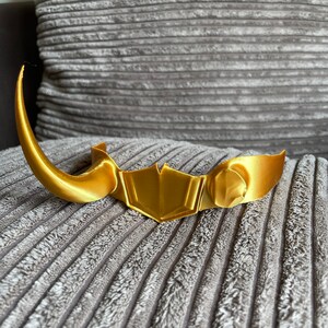 May include: A gold-colored plastic crown with a pointed horn on one side. The crown is on a gray textured surface.
