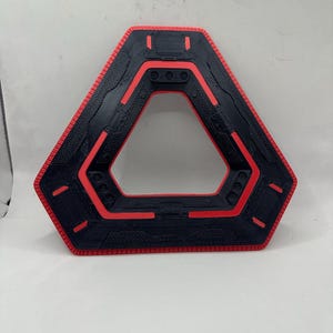May include: A black and red geometric object with a triangular centre. The outer edge is red, with a textured black surface and red accents. The object has a futuristic, tech-inspired design.