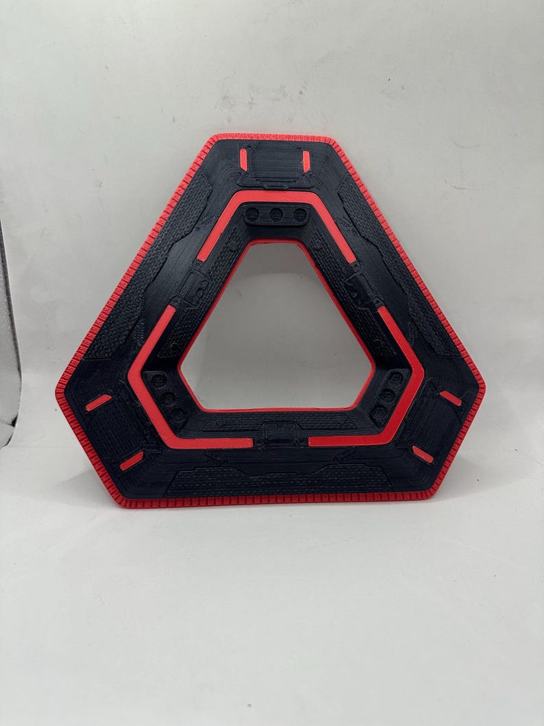 May include: A black and red geometric object with a triangular centre. The outer edge is red, with a textured black surface and red accents. The object has a futuristic, tech-inspired design.