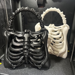 May include: Two skeleton-themed handbags, one black and one white, are displayed. The bags are designed to resemble rib cages, with handles that mimic vertebrae. The bags are on a dark surface, possibly inside a 3D printer.