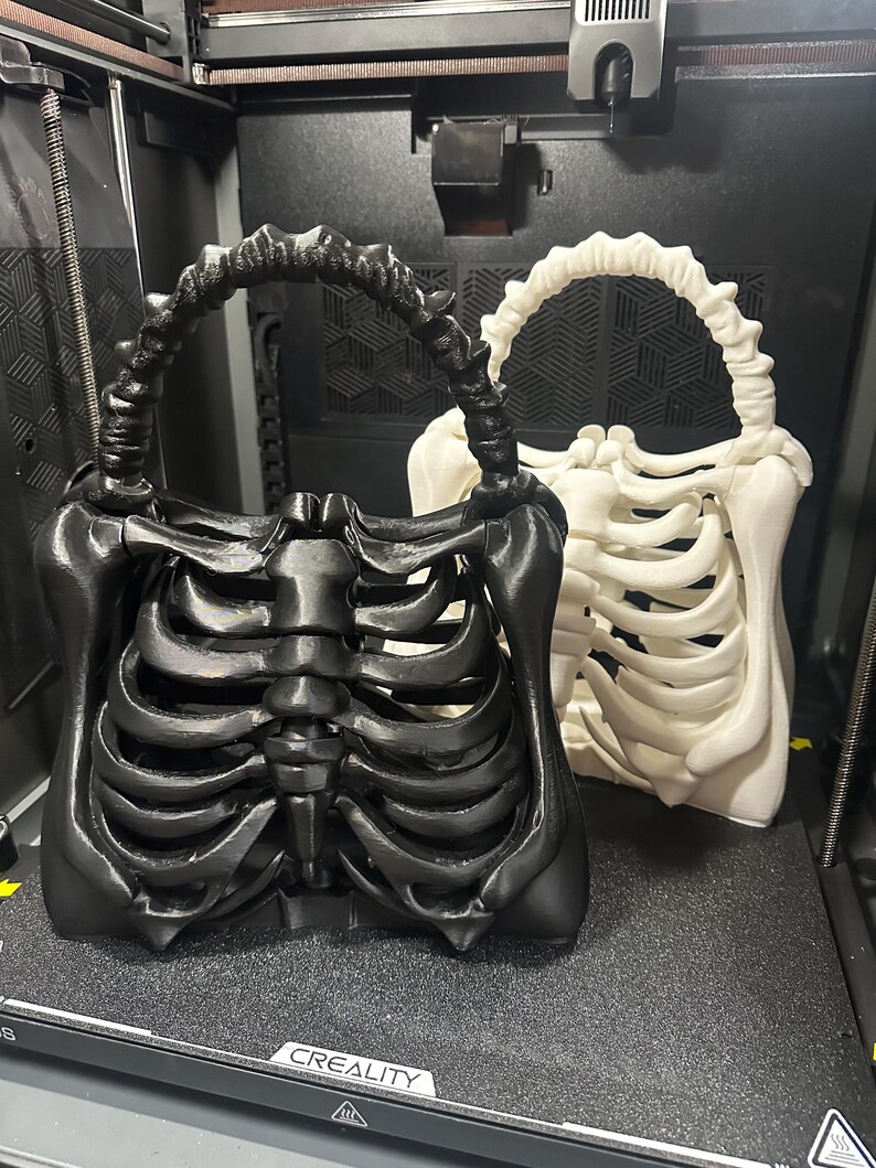 May include: Two skeleton-themed handbags, one black and one white, are displayed. The bags are designed to resemble rib cages, with handles that mimic vertebrae. The bags are on a dark surface, possibly inside a 3D printer.