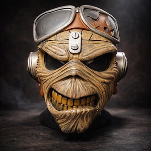 May include: A detailed, weathered mask with a menacing grin and yellow teeth. The mask features goggles, ear pieces, and a distressed finish, evoking a vintage or steampunk aesthetic. The color palette includes shades of brown, tan, and silver.