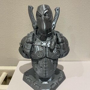 May include: A gray 3D printed bust of Deadpool, a superhero character, with a detailed design and a textured finish. The bust is posed with his arms crossed and his signature swords on his back.