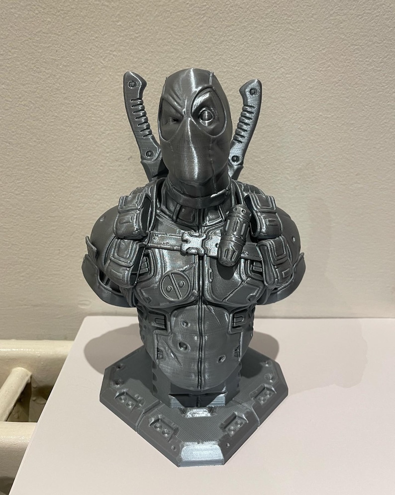 May include: A gray 3D printed bust of Deadpool, a superhero character, with a detailed design and a textured finish. The bust is posed with his arms crossed and his signature swords on his back.