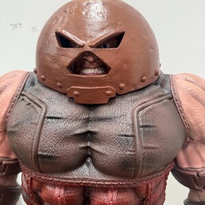 May include: A detailed figurine of a muscular character, featuring a brown helmet with eye slits and a menacing expression. The figure has a red and brown color scheme, with defined musculature and a textured appearance, showcasing a comic book or fantasy theme.