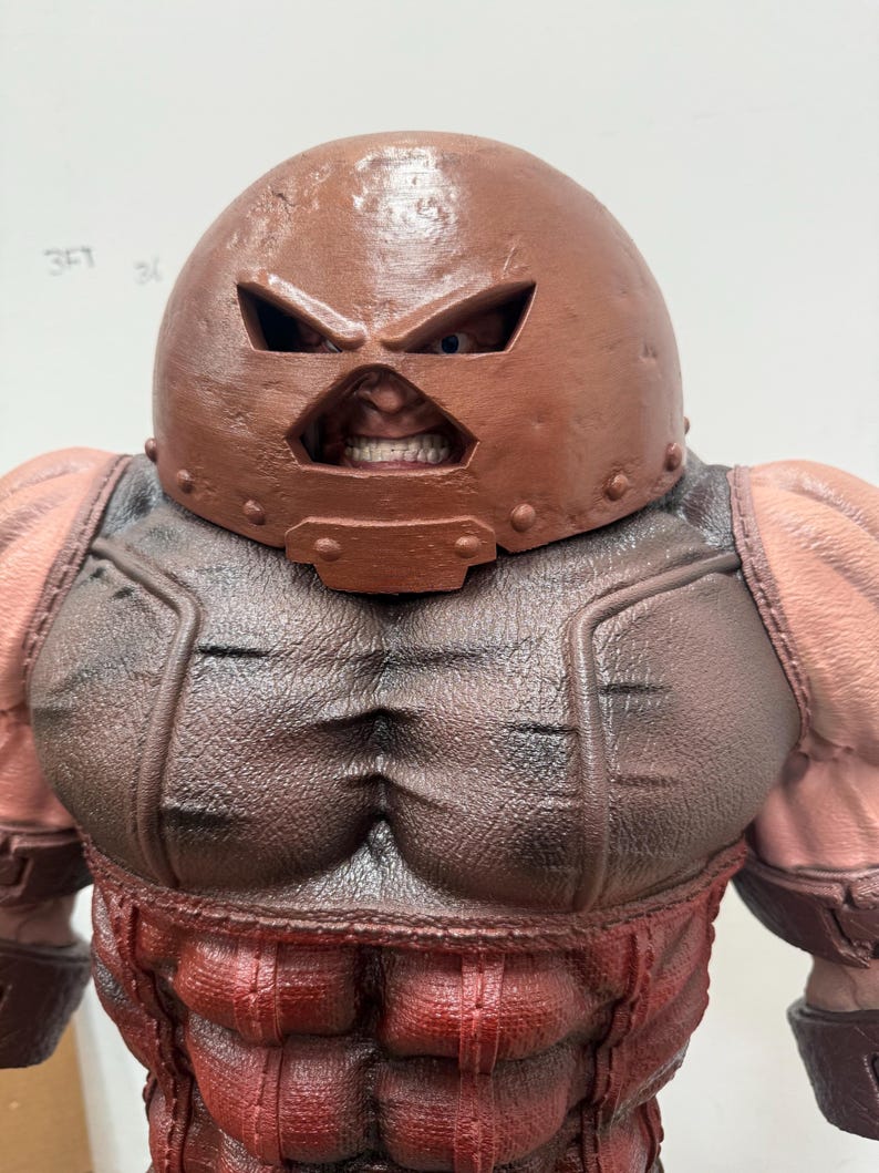 May include: A detailed figurine of a muscular character, featuring a brown helmet with eye slits and a menacing expression. The figure has a red and brown color scheme, with defined musculature and a textured appearance, showcasing a comic book or fantasy theme.
