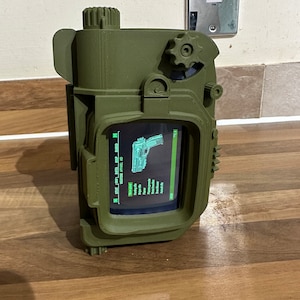 May include: A green 3D printed Pip-Boy replica with a screen displaying a green menu with the text "STAT  INV  DATA  MAP  AID  REPAIR  WHEEL  ID".