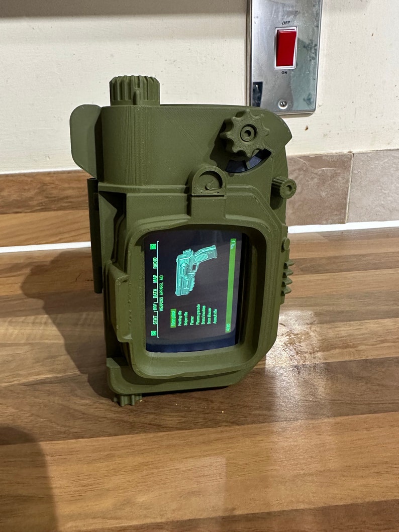 May include: A green 3D printed Pip-Boy replica with a screen displaying a green menu with the text "STAT  INV  DATA  MAP  AID  REPAIR  WHEEL  ID".