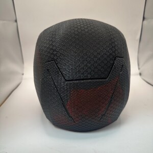 May include: A black, textured helmet with a geometric pattern. The helmet has a red accent on the lower back and a distinct angular design. The overall shape is rounded, suggesting a protective head covering.