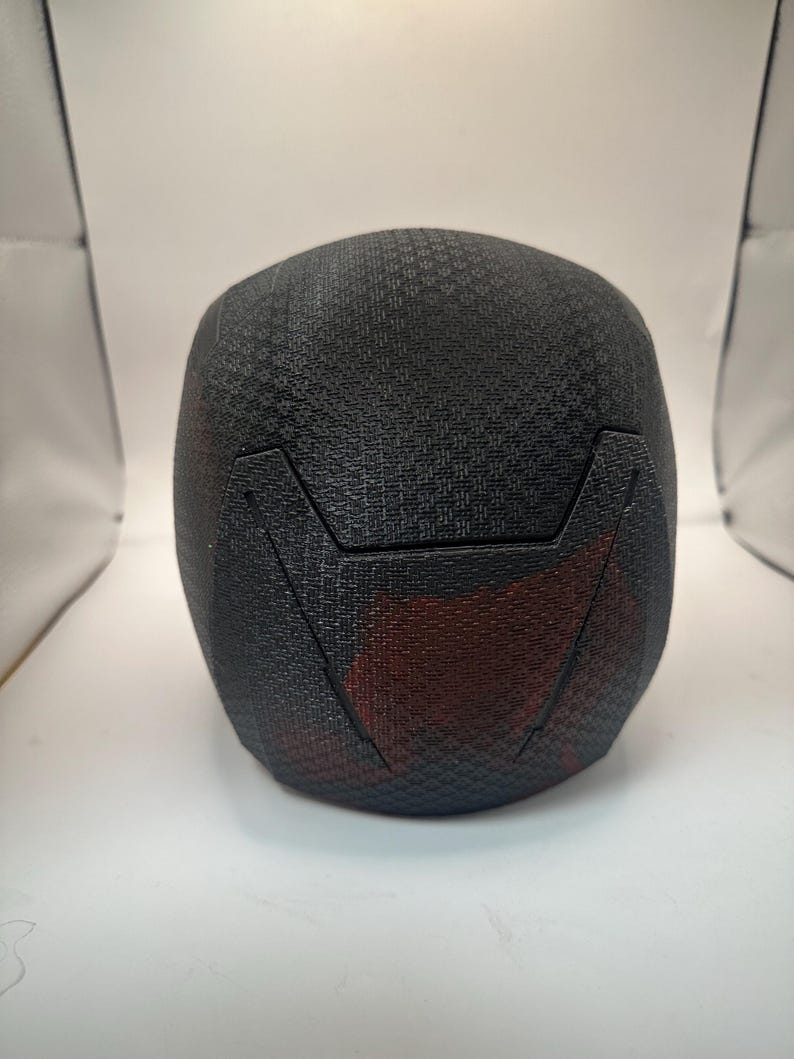 May include: A black, textured helmet with a geometric pattern. The helmet has a red accent on the lower back and a distinct angular design. The overall shape is rounded, suggesting a protective head covering.