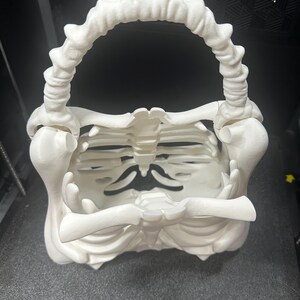 May include: A white, 3D-printed skeleton-themed basket with a handle. The basket is designed to resemble a rib cage, with a handle that arches over the top. The overall design is a unique and spooky decorative item.