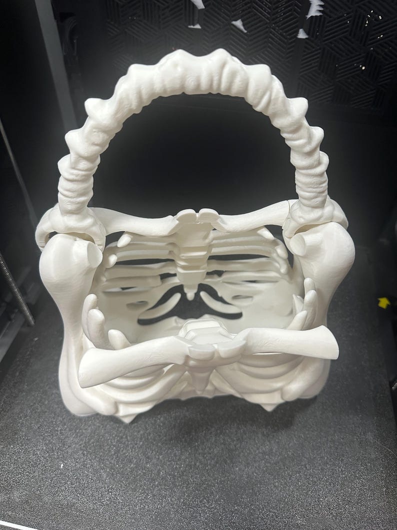 May include: A white, 3D-printed skeleton-themed basket with a handle. The basket is designed to resemble a rib cage, with a handle that arches over the top. The overall design is a unique and spooky decorative item.