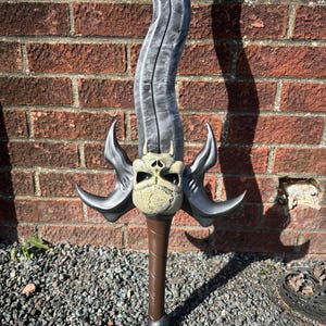 May include: A fantasy sword with a curved, gray blade and a detailed hilt. The hilt features a skull-like design with curved, gray accents. The handle is brown, and the base is dark gray. The sword is set against a red brick wall.