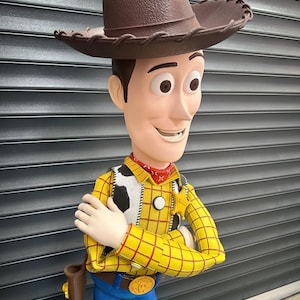 May include: A Woody action figure, wearing a brown cowboy hat, yellow plaid shirt, blue jeans, and a red neckerchief. The figure has a holster and a belt buckle with a gold emblem. The character's arms are crossed.